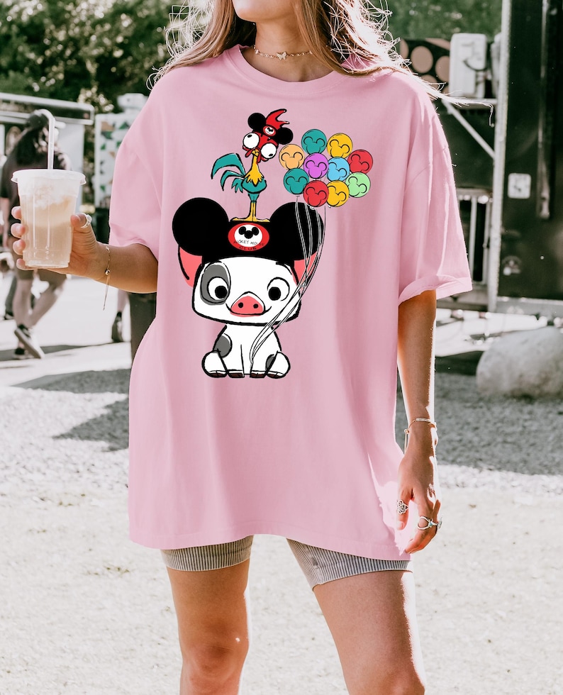 May include: A light pink t-shirt featuring a cartoon pig character with Mickey Mouse ears, holding colorful balloons. A cartoon rooster sits atop the pig's head. The shirt is a loose fit.