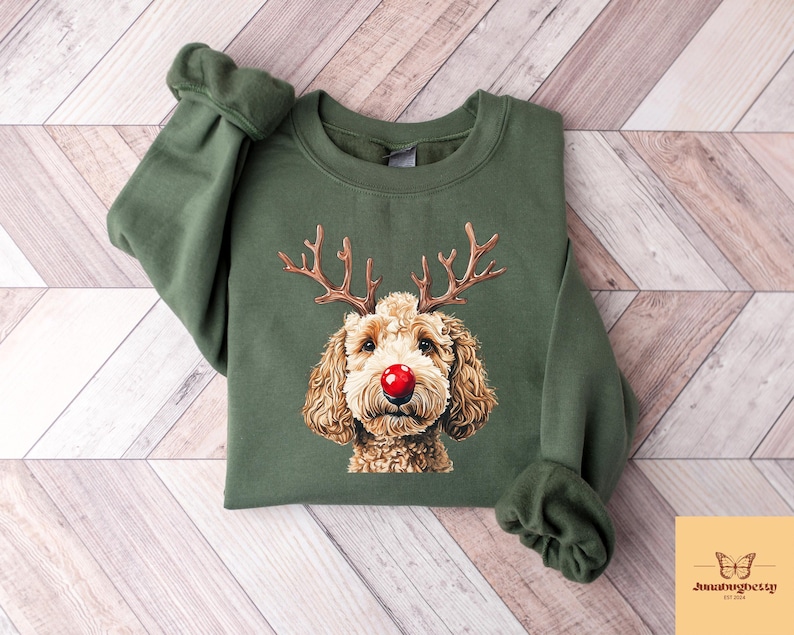 May include: Olive green sweatshirt featuring a graphic of a dog with reindeer antlers and a red nose. The dog has brown fur and is centered on the front of the long-sleeved top. The sweatshirt has a crew neck and ribbed cuffs.