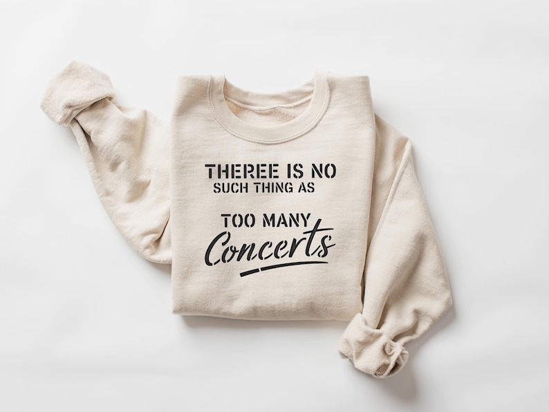 May include: Cream-colored sweatshirt with the text "THERE IS NO SUCH THING AS TOO MANY Concerts" in black. The sweatshirt is folded with the sleeves rolled up, and is set against a white background.