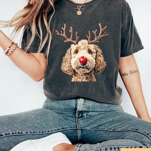 May include: Dark gray t-shirt featuring a dog with reindeer antlers and a red nose. The dog's face is the focal point, with detailed fur and a festive design. The shirt is a comfortable fit, perfect for casual wear.