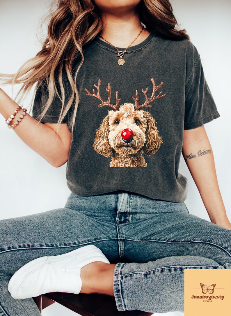 May include: Dark gray t-shirt featuring a dog with reindeer antlers and a red nose. The dog's face is the focal point, with detailed fur and a festive design. The shirt is a comfortable fit, perfect for casual wear.