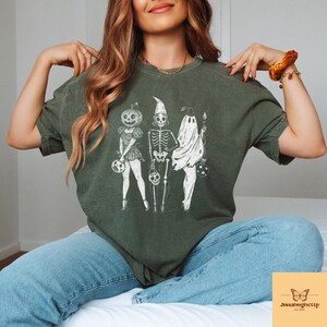 May include: Dark green t-shirt featuring a Halloween-themed graphic of three figures: a pumpkin-headed character, a skeleton, and a ghost. Each figure holds a Halloween treat bag. The shirt is paired with blue jeans.