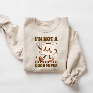 May include: Beige sweatshirt with the text "I'M NOT A GOOD WIPER" and a cartoon toilet paper roll character. The character has a smiling face, arms, legs, and red shoes. The design is in brown and red.