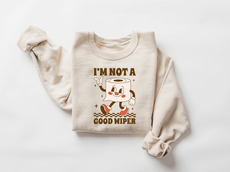 May include: Beige sweatshirt with the text "I'M NOT A GOOD WIPER" and a cartoon toilet paper roll character. The character has a smiling face, arms, legs, and red shoes. The design is in brown and red.