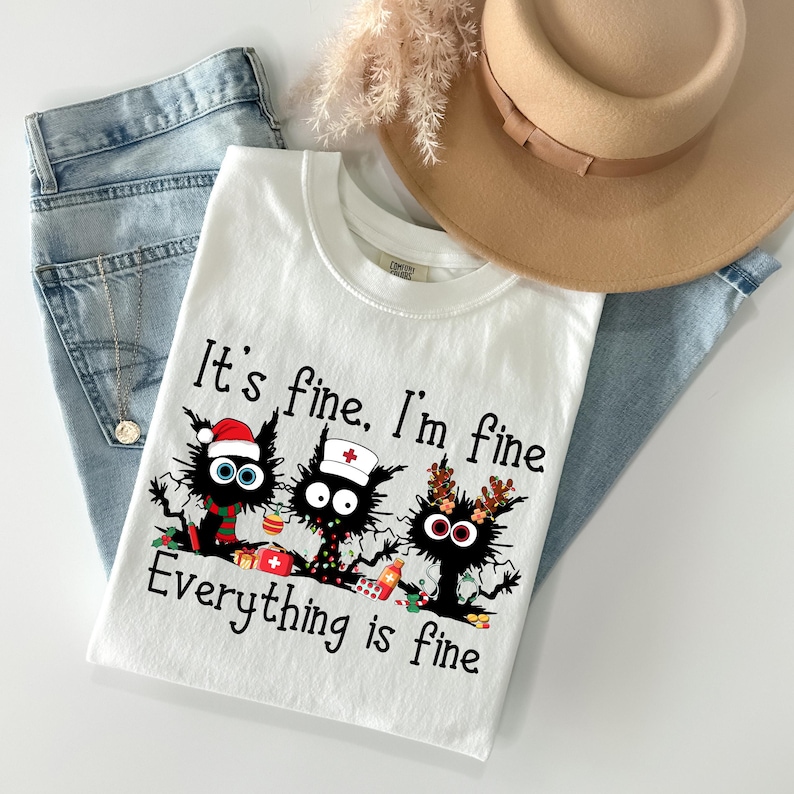 Może przedstawiać: Biała koszulka z napisem "It's fine, I'm fine, Everything is fine" i grafiką trzech czarnych kot&oacute;w w świątecznych akcesoriach. Koszulka jest zestawiona z niebieskimi dżinsami i beżowym kapeluszem.