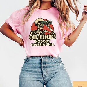 Puede incluir: Camiseta rosa claro con un dise&ntilde;o humor&iacute;stico de un esqueleto con prism&aacute;ticos dentro de un c&iacute;rculo rojo. El texto "OH LOOK! NOBODY GIVES A SHIT." est&aacute; impreso en negrita. Prenda de vestir informal.