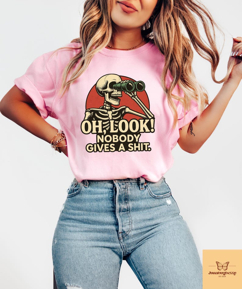 Puede incluir: Camiseta rosa claro con un dise&ntilde;o humor&iacute;stico de un esqueleto con prism&aacute;ticos dentro de un c&iacute;rculo rojo. El texto "OH LOOK! NOBODY GIVES A SHIT." est&aacute; impreso en negrita. Prenda de vestir informal.
