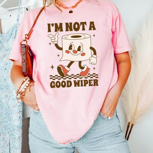 May include: Pink t-shirt with the text "I'M NOT A GOOD WIPER" and a cartoon toilet paper roll character. The character has a smiling face, arms, legs, and red shoes. The shirt is a light pink color.