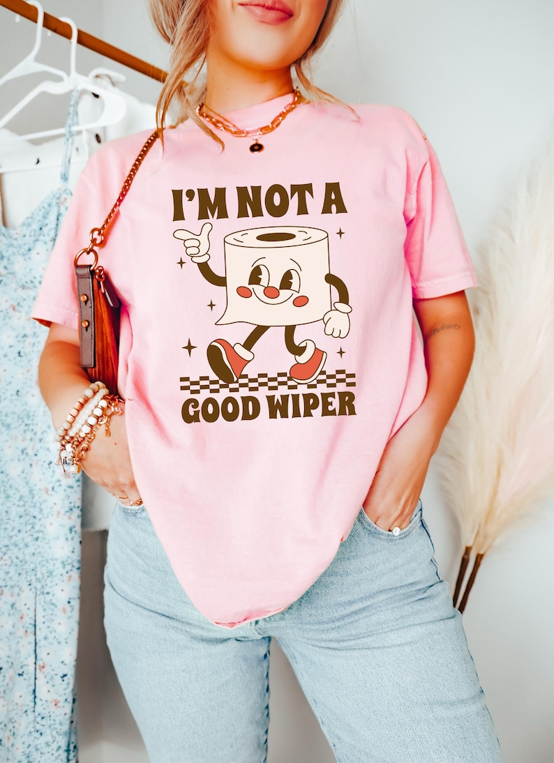 May include: Pink t-shirt with the text "I'M NOT A GOOD WIPER" and a cartoon toilet paper roll character. The character has a smiling face, arms, legs, and red shoes. The shirt is a light pink color.