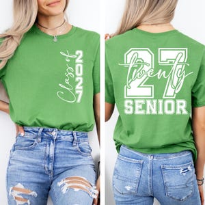 May include: A lime green t-shirt with white text. The front of the shirt says "Class of 2027" vertically. The back of the shirt says "27 Twenty Senior" in a white block design. The shirt is made of a soft material.