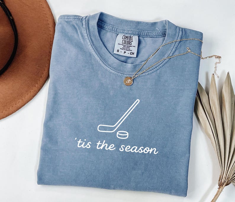 May include: A blue Comfort Colors t-shirt with a white hockey stick and puck graphic, and the phrase "'tis the season" printed below. A gold necklace with a pendant is draped across the shirt.