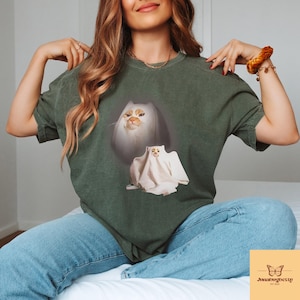 以下が含まれることがあります： ダークグリーンのTシャツ。気まぐれな猫のデザインが特徴です。グラフィックには、オレンジ色の模様と幽霊のコスチュームを着た白い猫が含まれています。このシャツは快適でカジュアルなスタイルです。