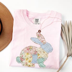 Puede incluir: Una camiseta rosa claro Comfort Colors con un dise&ntilde;o de conejo de retazos. El conejo est&aacute; hecho de telas florales, de cuadros y de encaje. Un delicado collar con un colgante descansa sobre la camisa. Un sombrero marr&oacute;n es visible en la esquina superior izquierda.