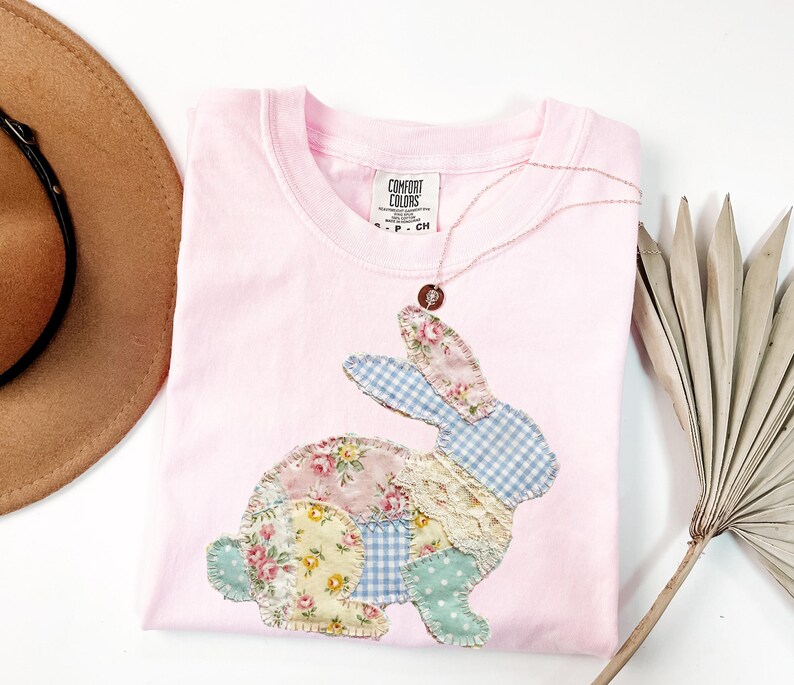 Puede incluir: Una camiseta rosa claro Comfort Colors con un dise&ntilde;o de conejo de retazos. El conejo est&aacute; hecho de telas florales, de cuadros y de encaje. Un delicado collar con un colgante descansa sobre la camisa. Un sombrero marr&oacute;n es visible en la esquina superior izquierda.