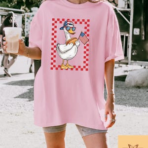 Op de afbeelding: Een roze oversized t-shirt met een afbeelding van een cartoon eend die een rood, wit en blauwe baseballpet draagt, een zonnebril en een hotdog en een Amerikaanse vlag vasthoudt. De eend staat op een rood-wit geruite achtergrond.