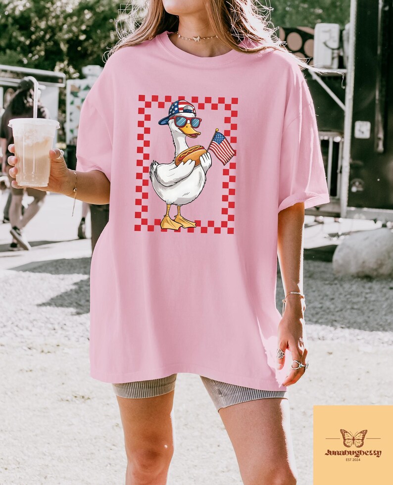 Op de afbeelding: Een roze oversized t-shirt met een afbeelding van een cartoon eend die een rood, wit en blauwe baseballpet draagt, een zonnebril en een hotdog en een Amerikaanse vlag vasthoudt. De eend staat op een rood-wit geruite achtergrond.