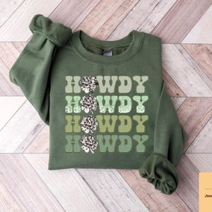 May include: Green sweatshirt with the text "Howdy" repeated four times in a gradient of green shades. Each "Howdy" is decorated with a white and green shamrock design.