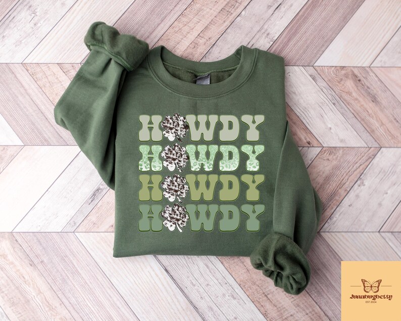 May include: Green sweatshirt with the text "Howdy" repeated four times in a gradient of green shades. Each "Howdy" is decorated with a white and green shamrock design.