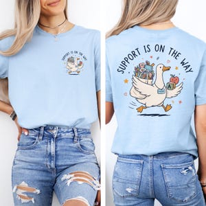 Op de afbeelding: Een lichtblauw t-shirt met een afbeelding van een witte eend die een tas met schoolspullen, een appel en een blok met de letters AI draagt. De tekst "SUPPORT IS ON THE WAY" is in het zwart boven de eend gedrukt op zowel de voor- als achterkant.