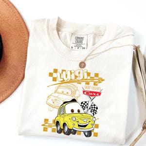 May include: Cream-coloured t-shirt featuring a yellow cartoon car named Luigi from the movie Cars. The design includes the name "Luigi", the text "EST 2006 Cars", and checkered flags. A gold necklace and a brown hat are also visible.