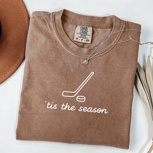 May include: A brown t-shirt with the text "'tis the season" and a hockey stick and puck graphic. The shirt has a crew neck and short sleeves. A silver necklace with a pendant is draped across the shirt.