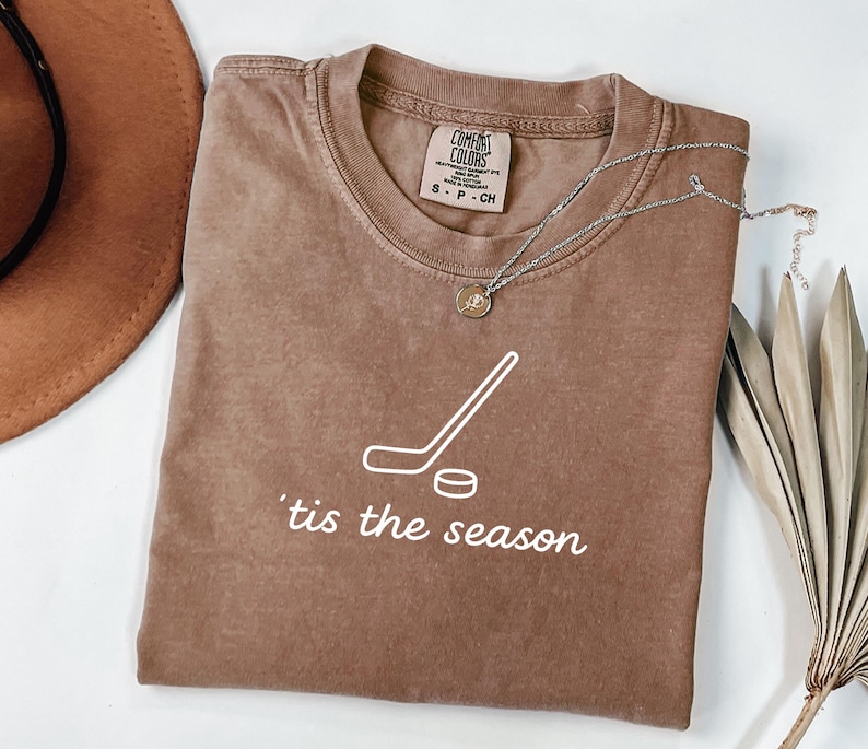 May include: A brown t-shirt with the text "'tis the season" and a hockey stick and puck graphic. The shirt has a crew neck and short sleeves. A silver necklace with a pendant is draped across the shirt.
