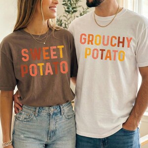 May include: A dark brown t-shirt with the text SWEET POTATO in orange, yellow, and pink, and a white t-shirt with the text GROUCHY POTATO in orange, yellow, and pink. Both shirts feature short sleeves and a crew neckline, worn with blue denim jeans.