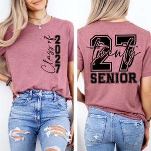 May include: A mauve-colored t-shirt with the text "Class of 2027" printed vertically on the front. The back of the shirt features the text "27 Twenty Senior" in a black, bold font. The shirt is paired with blue jeans.