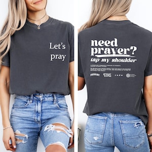 Puede incluir: Camiseta gris oscuro con el texto "Let's pray" en la parte delantera. La parte trasera de la camiseta tiene el texto "need prayer? tap my shoulder" en blanco. La camiseta se combina con vaqueros azules.
