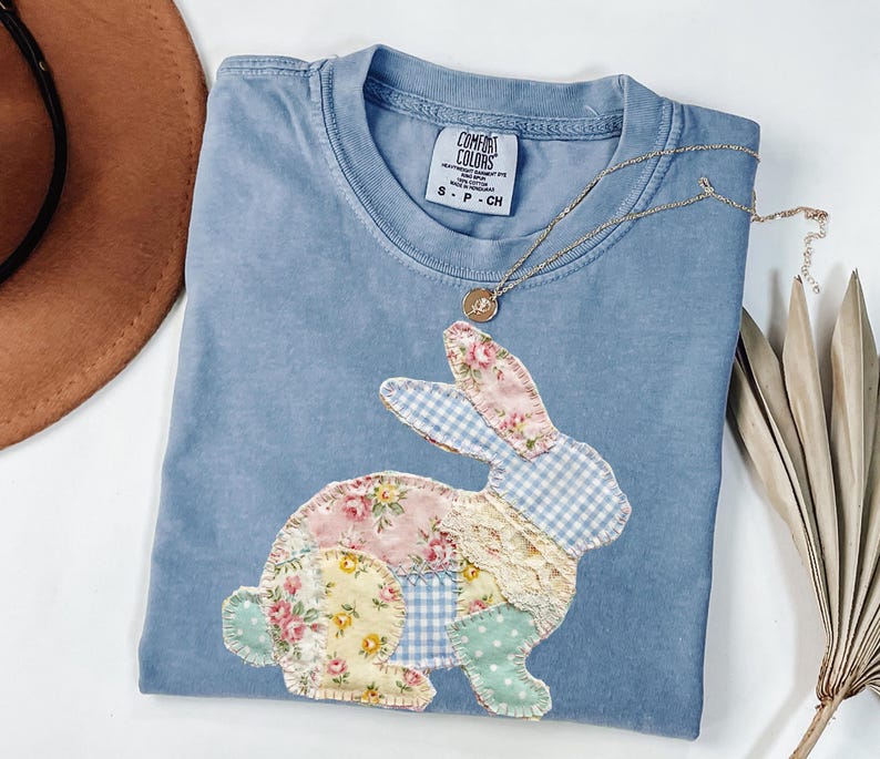 Puede incluir: Una camiseta Comfort Colors azul claro con un dise&ntilde;o de conejito de patchwork. El conejito est&aacute; hecho de telas florales, de cuadros y de encaje. Un collar dorado con un colgante est&aacute; drapeado sobre la camiseta. Un sombrero marr&oacute;n est&aacute; en el fondo.