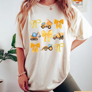 May include: A cream-coloured t-shirt with a design of yellow bows and construction vehicles. The vehicles include a dumper truck, excavator, cement mixer, and a front-end loader. The shirt has a relaxed fit and short sleeves.