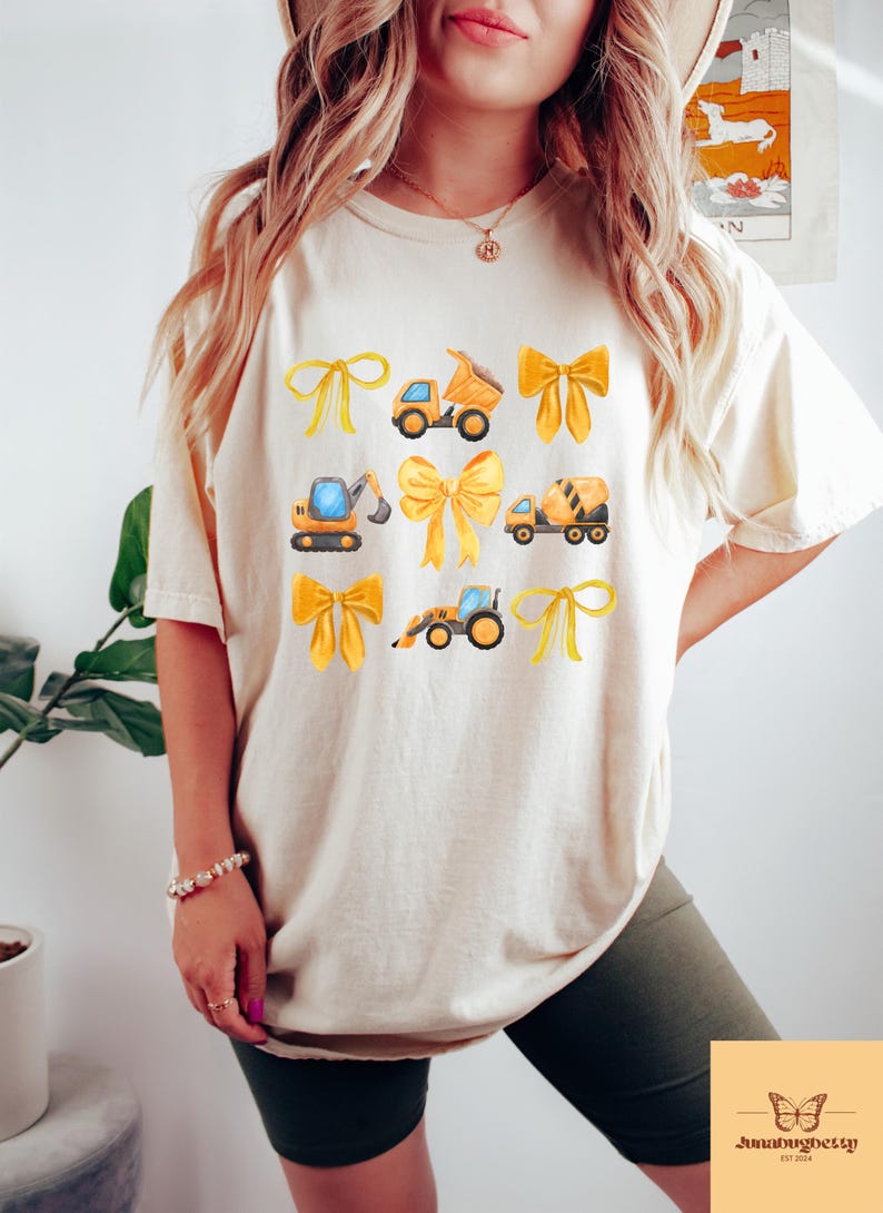 May include: A cream-coloured t-shirt with a design of yellow bows and construction vehicles. The vehicles include a dumper truck, excavator, cement mixer, and a front-end loader. The shirt has a relaxed fit and short sleeves.