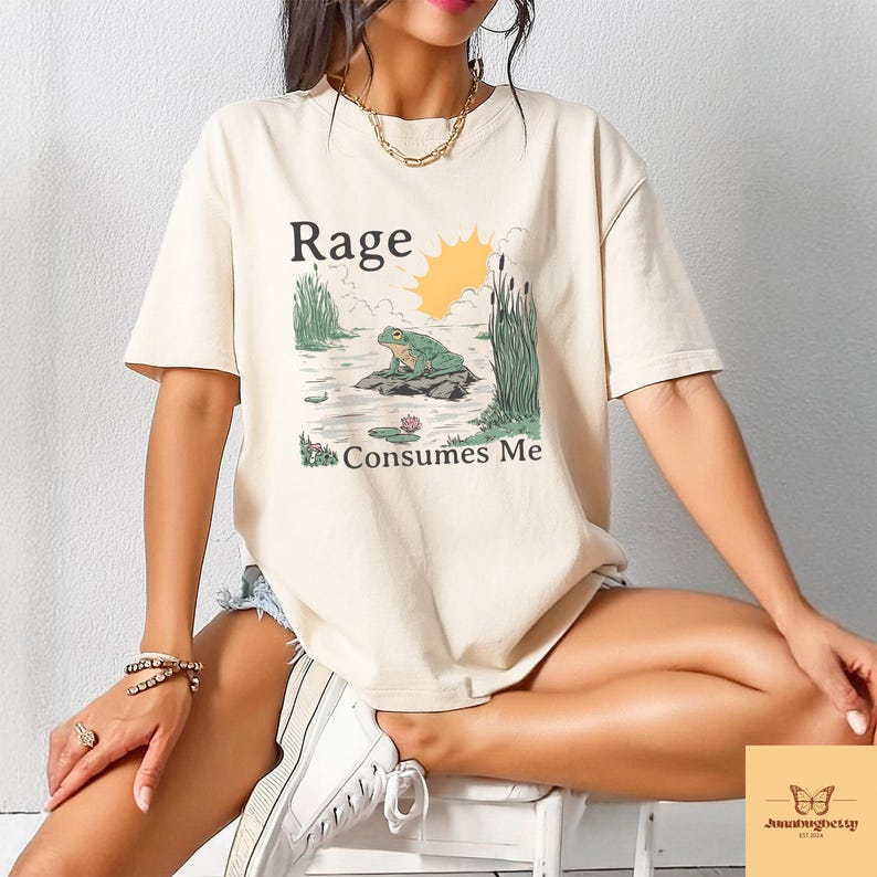 May include: Cream-colored t-shirt featuring a graphic of a frog on a rock in a pond, with the text "Rage Consumes Me." The design includes a sun, reeds, and lily pads. The shirt has short sleeves.