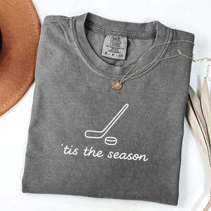 May include: A gray Comfort Colors t-shirt with a white hockey stick and puck graphic, and the phrase "'tis the season" in white script. A gold necklace with a pendant is draped on the shirt.