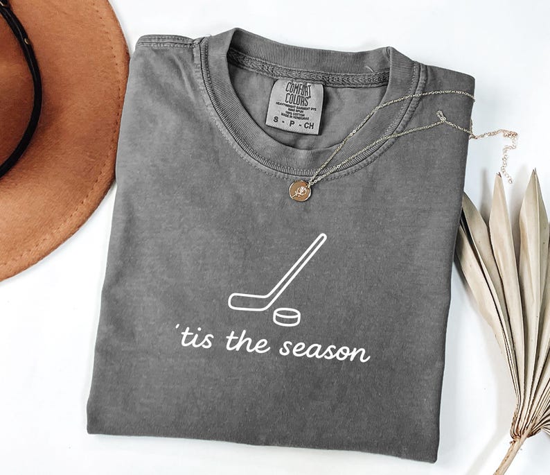 May include: A gray Comfort Colors t-shirt with a white hockey stick and puck graphic, and the phrase "'tis the season" in white script. A gold necklace with a pendant is draped on the shirt.