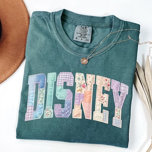 May include: A dark teal short-sleeve t-shirt featuring the word DISNEY in large, block-style letters. Each letter is filled with a patchwork of pastel-coloured fabrics in floral, plaid, and gingham patterns. The shirt has a Comfort Colors label.