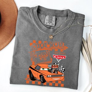 May include: Grey t-shirt with an orange cartoon car design, featuring the text "Shoot Rod" and "Cars". The design includes checkered flags and a vintage car illustration. The shirt is a Comfort Colors brand.