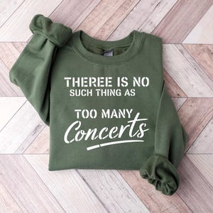 May include: An olive green sweatshirt with the text "THERE IS NO SUCH THING AS TOO MANY Concerts" in white. The sweatshirt is folded on a light wood grain surface.