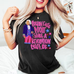 May include: Black t-shirt featuring a cartoon illustration of a historical figure in a blue coat and pink striped pants. The shirt has the text "Feelin' Cute Might Start A Revolution Later, IDK" in pink and blue.