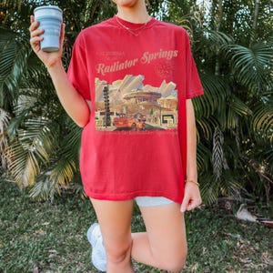 以下が含まれることがあります： カリフォルニア州ラジエーター・スプリングスのグラフィックがプリントされた赤いTシャツ。デザインには、ヴィンテージカー、建物、テキスト「Radiator Springs」と「California Est. 2001」が含まれています。このシャツはクルーネックで半袖です。