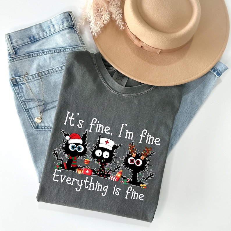 Może przedstawiać: Ciemnoszara koszulka z napisem "It's fine, I'm fine, Everything is fine" i grafiką trzech czarnych kot&oacute;w w świątecznych akcesoriach. Koszulka jest zestawiona z jasnoniebieskimi dżinsami i beżowym kapeluszem.