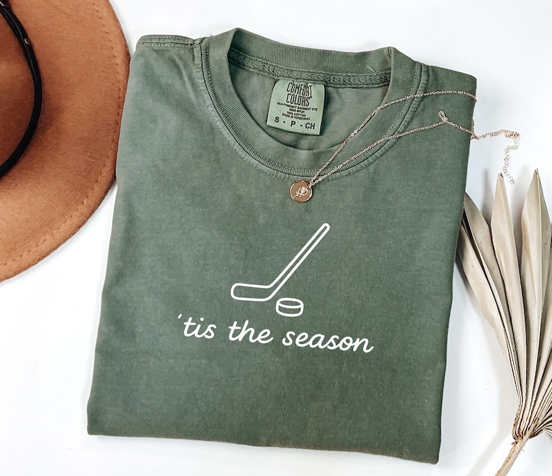 May include: An olive green t-shirt with a white hockey stick and puck graphic, and the phrase "'tis the season" in white. A gold necklace with a pendant is draped across the shirt. A brown hat and dried palm leaf are in the background.