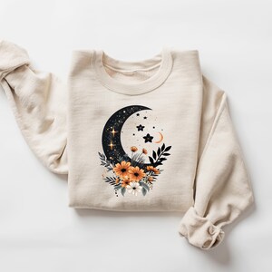 May include: Cream-colored sweatshirt featuring a celestial design. A black crescent moon with stars and a floral arrangement of orange and white flowers is printed on the front. The sleeves are rolled up.