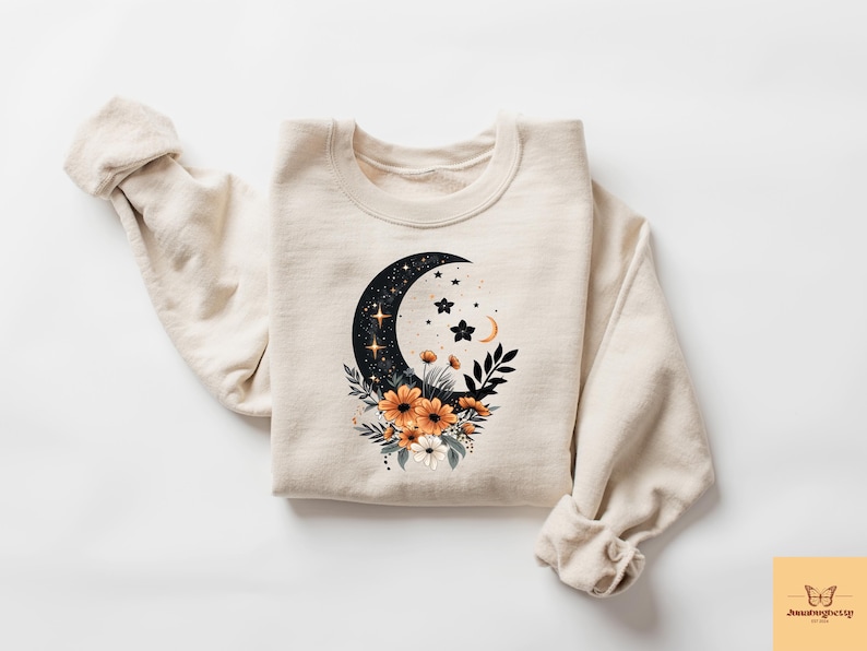 May include: Cream-colored sweatshirt featuring a celestial design. A black crescent moon with stars and a floral arrangement of orange and white flowers is printed on the front. The sleeves are rolled up.