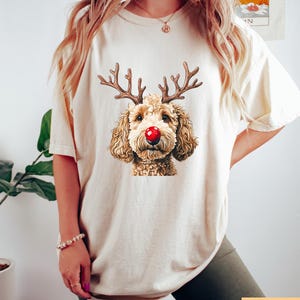 May include: Cream-colored t-shirt featuring a graphic of a dog with reindeer antlers and a red nose. The dog has brown fur and is centered on the shirt. The shirt is a casual style, suitable for everyday wear.