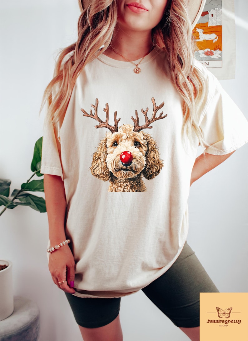 May include: Cream-colored t-shirt featuring a graphic of a dog with reindeer antlers and a red nose. The dog has brown fur and is centered on the shirt. The shirt is a casual style, suitable for everyday wear.