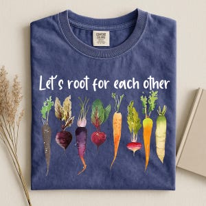 May include: A navy blue crew neck t-shirt featuring the white text Let's root for each other above a colourful watercolour illustration of eight different root vegetables including carrots, beets, and radishes in shades of purple, red, orange, and green.
