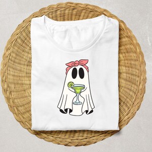 Pu&ograve; includere: Maglietta bianca con un fantasma dei cartoni animati che indossa un bandana rosa e tiene un bicchiere di margarita. Il fantasma ha uno spicchio di lime nel suo bicchiere.