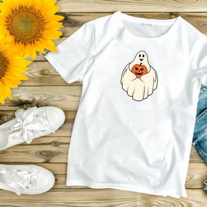 Pu&ograve; includere: Maglietta bianca con un fantasma che tiene una zucca. Il fantasma sorride e la zucca ha una faccia spettrale. La maglietta &egrave; su una superficie di legno con girasoli e jeans.