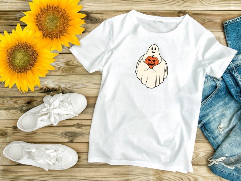 Pu&ograve; includere: Maglietta bianca con un fantasma che tiene una zucca. Il fantasma sorride e la zucca ha una faccia spettrale. La maglietta &egrave; su una superficie di legno con girasoli e jeans.